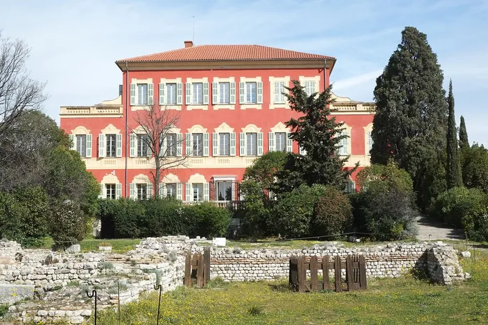The red-ochre Villa des Arènes, home of the Musée Matisse, behind cypresses and lawns on the Cimiez hill, with low Roman ruins in the foreground.