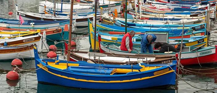 Traditional wooden fishing boats painted blue, red, yellow and white moored close together in Port Lympia, Nice, with two men working on one of them.