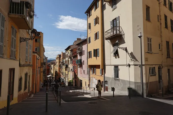 A narrow sunlit lane in Vieux-Nice lined with ochre and pale-yellow shuttered buildings with wrought-iron balconies, a pedestrian walking away down the cobbled street under a blue sky.