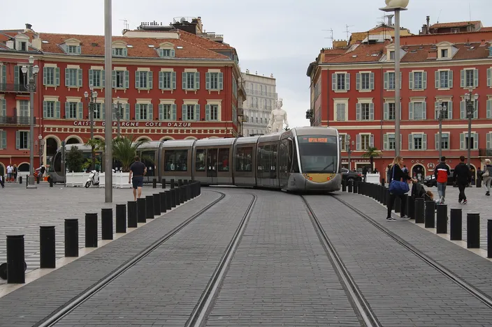 A silver Citadis tram glides across the chequered paving of Place Masséna in Nice, between the red-ochre arcaded buildings, with pedestrians and black bollards either side of the tracks.
