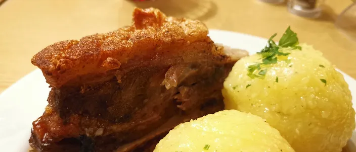 A thick slab of roast pork shoulder with a blistered golden crackling crust beside two pale potato dumplings garnished with parsley