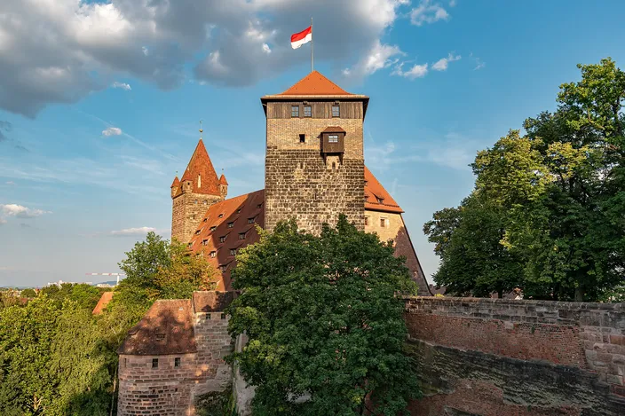 A stone tower and red roofs of Nuremberg's Imperial Castle above its walls and trees, a red-and-white flag flying against a blue sky