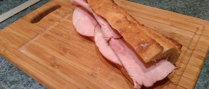A split length of baguette on a wooden board with slices of pale pink cooked ham laid inside, the classic jambon-beurre.