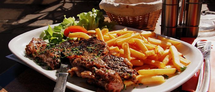 A seared, herb-topped steak beside a heap of golden fries with a small salad garnish, on a sunlit bistro table.