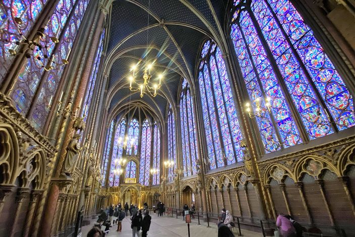 The upper chapel of the Sainte-Chapelle, tall stained-glass windows in deep blue and red running from waist height to a star-painted vault, with a scatter of visitors on the floor below