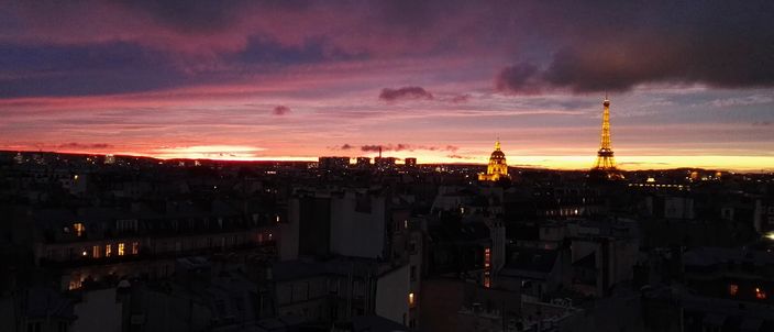 Paris rooftops at dusk beneath a red and violet sunset, the Eiffel Tower lit gold on the horizon at right and the gilded dome of Les Invalides glowing beside it