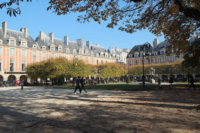 Place des Vosges in autumn: identical brick-and-stone pavilions with steep slate roofs above a continuous ground-floor arcade, two people crossing the sandy ground under yellowing trees