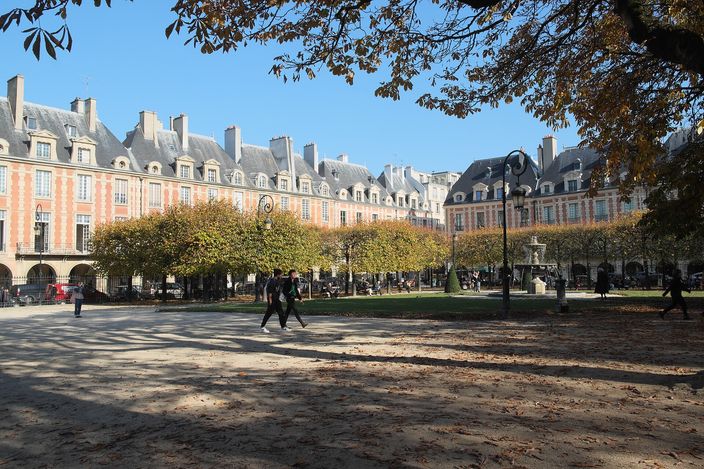 Place des Vosges in autumn: identical brick-and-stone pavilions with steep slate roofs above a continuous ground-floor arcade, two people crossing the sandy ground under yellowing trees