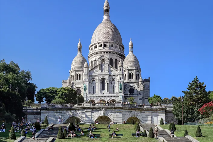 The Sacre-Coeur basilica in white stone against a cloudless blue sky, its dome and two flanking towers above an arcaded terrace, with people sitting on the clipped lawns and stairs below