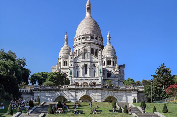 The Sacre-Coeur basilica in white stone against a cloudless blue sky, its dome and two flanking towers above an arcaded terrace, with people sitting on the clipped lawns and stairs below