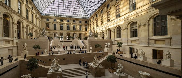 The glass-roofed Cour Marly inside the Louvre: French sculptures on stone plinths stepped down a terraced courtyard beneath a vast pitched skylight, visitors small among them