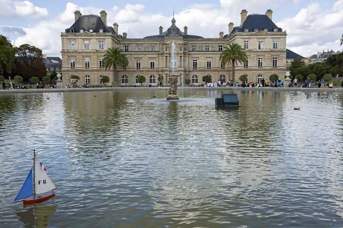 The Luxembourg Palace across the Grand Bassin in the Jardin du Luxembourg, a small model sailboat with a French flag on its sail in the foreground water, palms and clipped trees along the terrace and people sitting round the basin