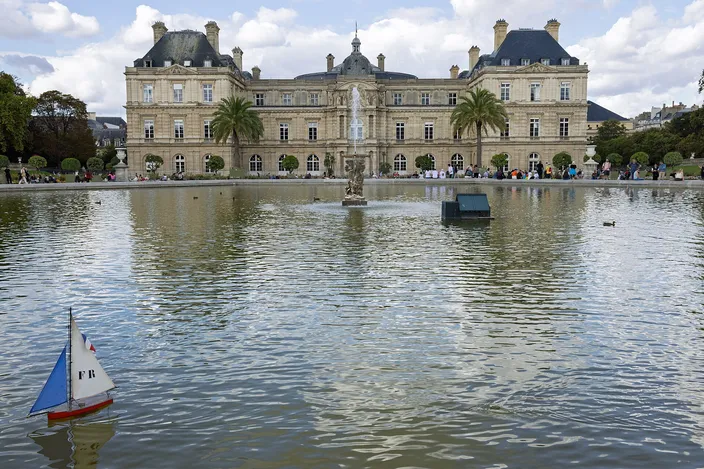 The Luxembourg Palace across the Grand Bassin in the Jardin du Luxembourg, a small model sailboat with a French flag on its sail in the foreground water, palms and clipped trees along the terrace and people sitting round the basin