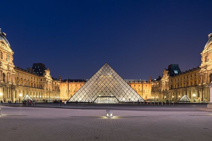 The Cour Napoléon of the Louvre at blue hour, the glass pyramid lit from within at the centre of the empty courtyard with the palace wings floodlit gold on either side under a deep blue sky