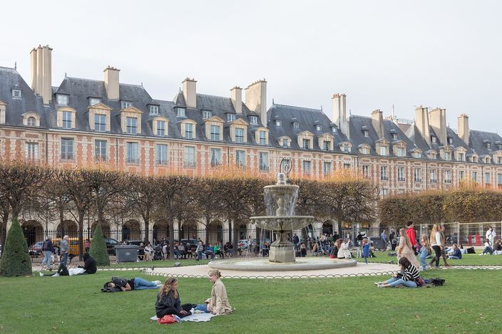The Place des Vosges in autumn, red-brick and pale-stone pavilions with steep slate roofs and tall chimneys standing to one design along two sides, a stone fountain playing in the middle and people lying on the lawn in front of it