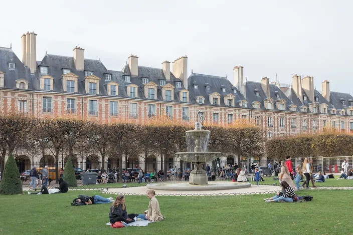 The Place des Vosges in autumn, red-brick and pale-stone pavilions with steep slate roofs and tall chimneys standing to one design along two sides, a stone fountain playing in the middle and people lying on the lawn in front of it