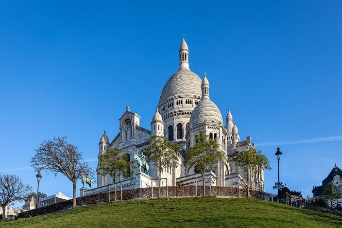 The Basilica of Sacré-Cœur seen from the south-east, its white domes and campanile bright against a cloudless deep-blue sky, standing above a green grass slope with bare young trees and bronze equestrian statues on the terrace below