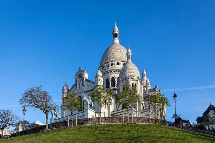 The Basilica of Sacré-Cœur seen from the south-east, its white domes and campanile bright against a cloudless deep-blue sky, standing above a green grass slope with bare young trees and bronze equestrian statues on the terrace below