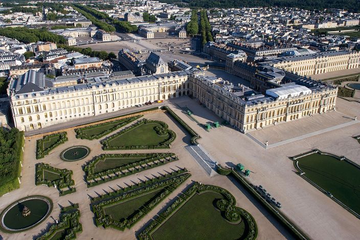 An aerial view of the Palace of Versailles, its long garden front and pavilions in pale stone with the clipped scrollwork parterres and a fountain basin laid out below, the forecourt and the roofs of the town stretching away behind