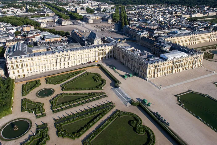 An aerial view of the Palace of Versailles, its long garden front and pavilions in pale stone with the clipped scrollwork parterres and a fountain basin laid out below, the forecourt and the roofs of the town stretching away behind