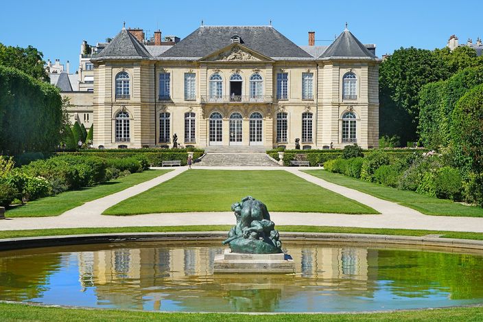 The garden front of the Hôtel Biron at the Musée Rodin, a wide eighteenth-century stone house with tall windows and slate roofs at the head of a lawn, gravel walks and clipped hedges on either side and a dark bronze group standing in a still round pond in the foreground