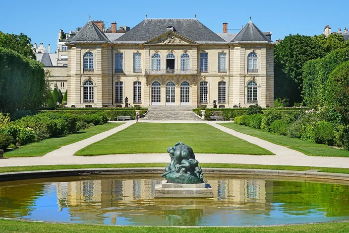 The garden front of the Hôtel Biron at the Musée Rodin, a wide eighteenth-century stone house with tall windows and slate roofs at the head of a lawn, gravel walks and clipped hedges on either side and a dark bronze group standing in a still round pond in the foreground