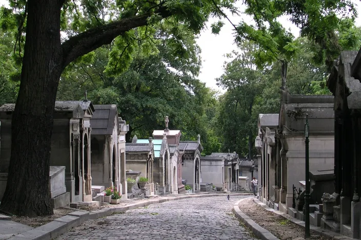 A cobbled lane in Père Lachaise cemetery running away between two close-packed rows of stone tombs built as miniature chapels with pointed gables, a large tree leaning over the road on the left and dense green trees closing the view ahead