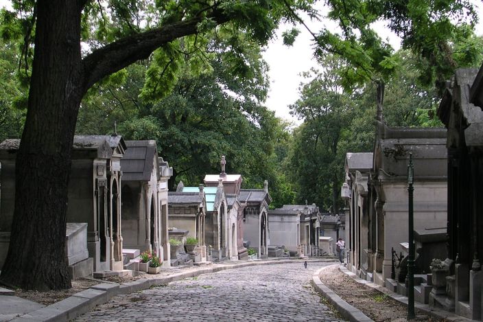 A cobbled lane in Père Lachaise cemetery running away between two close-packed rows of stone tombs built as miniature chapels with pointed gables, a large tree leaning over the road on the left and dense green trees closing the view ahead