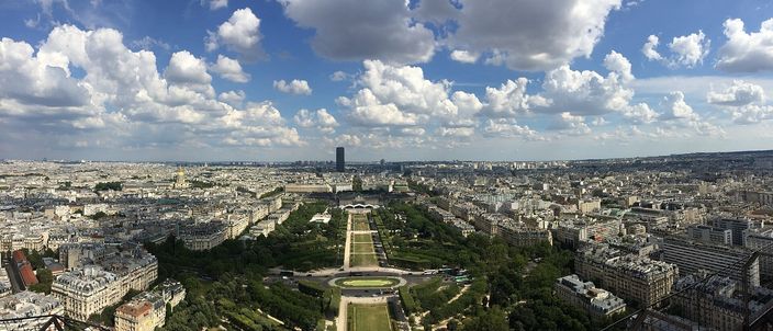 Paris seen from high on the Eiffel Tower, the long green lawn of the Champ de Mars running away to the École Militaire, dense pale rooftops on both sides and the dark Tour Montparnasse on the skyline under tall summer cloud