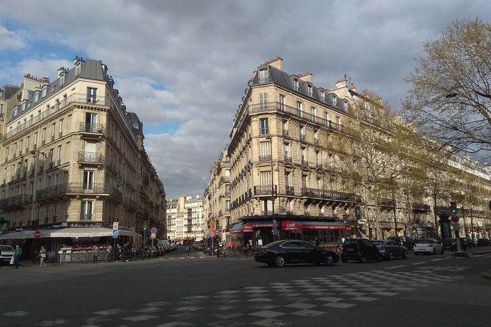 A corner of Haussmann apartment blocks with wrought-iron balconies and a café at street level, where Rue Dante meets Boulevard Saint-Germain, cars waiting at the crossing