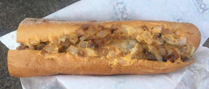 A Philadelphia cheesesteak on a long soft roll, filled with chopped griddled beef, onions and melted cheese sauce, resting on white paper.