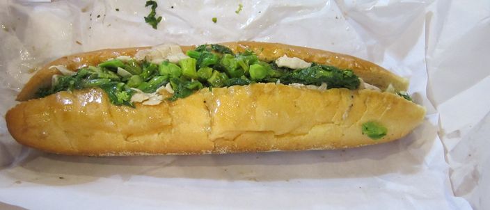 A Philadelphia roast pork sandwich on a long roll, piled with sliced roast pork and sauteed broccoli rabe, in its paper wrap.