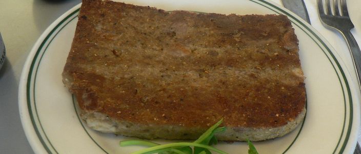 A thick square slice of pan-fried scrapple with a browned, crisp crust, served on a plate with a fork and a sprig of parsley.