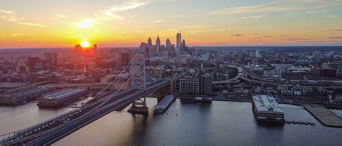 Aerial view at sunset of the Benjamin Franklin Bridge spanning the Delaware River, with the Center City Philadelphia skyline and its cluster of glass towers catching the last light behind it