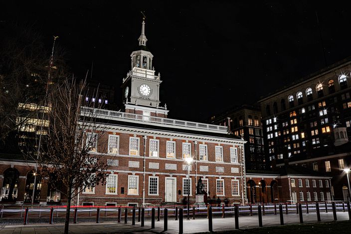 Independence Hall floodlit at night: a two-storey red-brick Georgian building with white-framed sash windows and a white clock steeple, seen head-on across an empty railed plaza with modern office blocks lit behind it