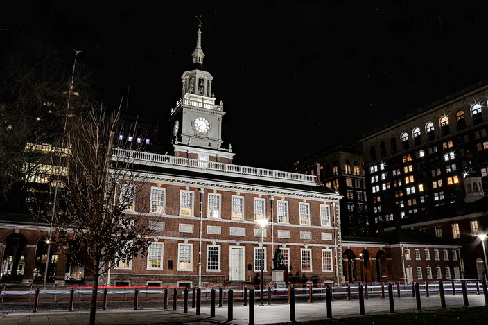 Independence Hall floodlit at night: a two-story red-brick Georgian building with white-framed sash windows and a white clock steeple, seen head-on across an empty railed plaza with modern office blocks lit behind it