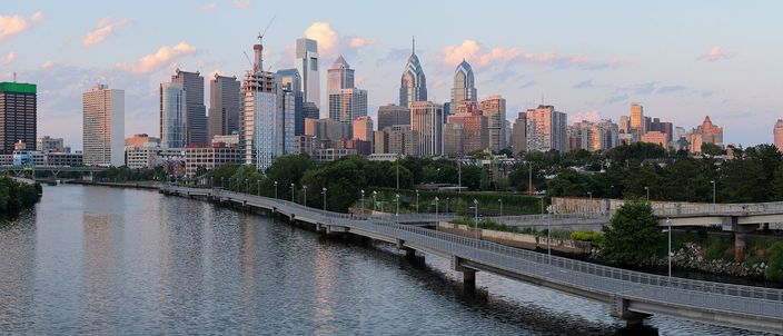The Schuylkill River in golden evening light seen from a bridge, with the riverside walkway and expressway running along the near bank and the Center City Philadelphia skyline of glass and stone towers rising behind the trees