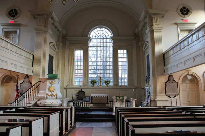 The interior of a Georgian church looking towards the altar: white-painted columns and galleries either side, a tall arched Palladian window filling the east wall behind a plain communion table, a raised wineglass pulpit with a carved crest at left, and rows of boxed wooden pews in the foreground