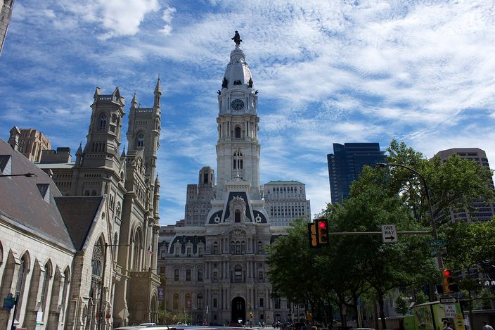 Philadelphia City Hall seen from a street corner under a blue sky with high cloud: a white masonry building with a mansard roof and an ornate tower rising above it, topped by a statue on a lantern, with the Gothic turrets of the Masonic Temple at left and modern glass towers and street trees at right
