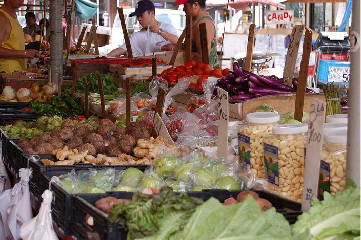An open-air market stall under a striped awning: crates and boxes of vegetables — cabbages, tomatoes, aubergines, ginger root, spring onions — banked up on trestles along a pavement, with handwritten price cards among them and stallholders working behind the counter