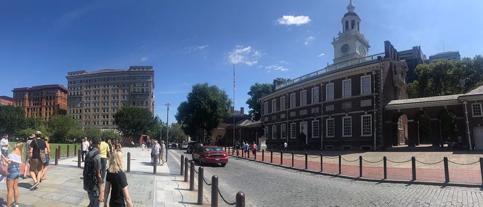 A wide daylight view along Chestnut Street with Independence Hall on the right: a two-storey red-brick Georgian building with white sash windows and a white clock steeple, fronted by a low black railing and brick paving, with pedestrians on the pavement at left and a mid-century office block and trees beyond