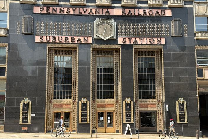 Art Deco black granite facade with gilded 'Pennsylvania Railroad Suburban Station' lettering above the entrance, two cyclists passing on the sidewalk