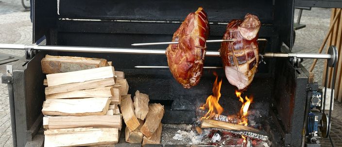 Two whole Prague hams on the bone roasting on a horizontal spit over an open wood fire at a street stall