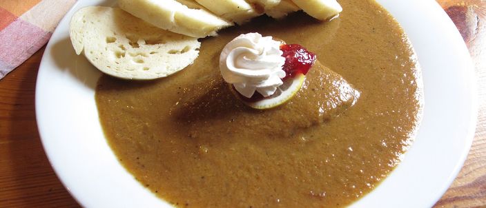 A plate of svíčková: beef under a thick pale sauce, a fan of sliced bread dumplings, topped with a swirl of whipped cream and a spoon of cranberry