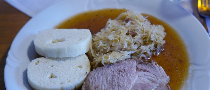 A plate of vepřo knedlo zelo: two slices of roast pork with gravy, two discs of white bread dumpling, and a mound of braised sauerkraut
