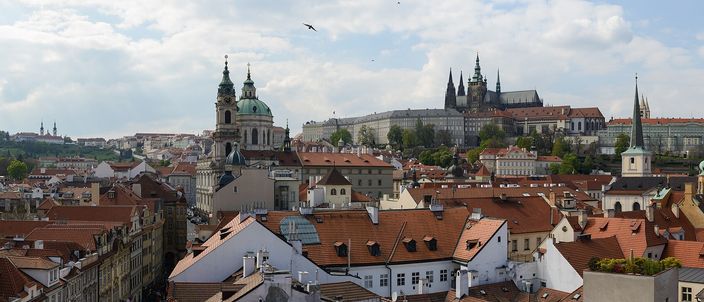 Panorama across the red-tiled roofs of Malá Strana to the Gothic spires of St Vitus Cathedral and Prague Castle on the hill, with the green dome of St Nicholas Church at left