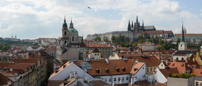 Panorama across the red-tiled roofs of Malá Strana to the Gothic spires of St Vitus Cathedral and Prague Castle on the hill, with the green dome of St Nicholas Church at left