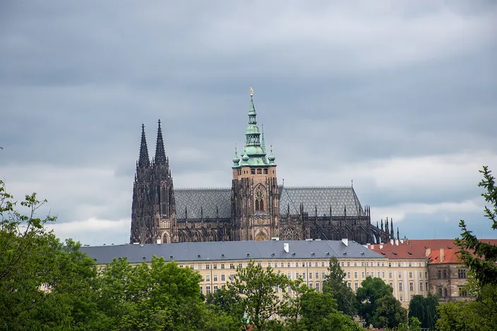 A gothic cathedral with two openwork stone spires at its west end and a taller square tower carrying a green copper cupola and a gold finial, its nave roof patterned in gray diamonds; below it a long pale palace wing with regular rows of windows and a gray roof, and leafy green trees filling the lower corners, under an overcast sky.