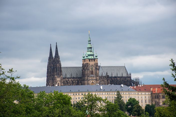 A gothic cathedral with two openwork stone spires at its west end and a taller square tower carrying a green copper cupola and a gold finial, its nave roof patterned in grey diamonds; below it a long pale palace wing with regular rows of windows and a grey roof, and leafy green trees filling the lower corners, under an overcast sky.