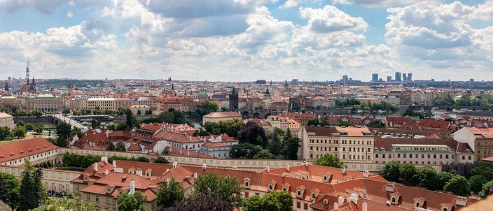 Red-tiled rooftops with dormer windows fill the foreground and fall away to a river crossed by several bridges; a long stone bridge with dark towers stands at the centre, and beyond it a dense city of spires, domes and pale facades runs to the horizon under white cumulus cloud.